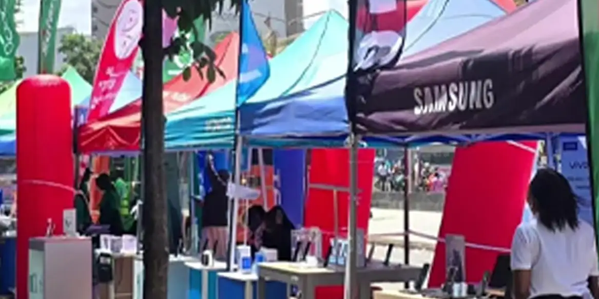 Row of colorful vendor booths at an outdoor expo with a Samsung tent in the foreground, people browsing displays.